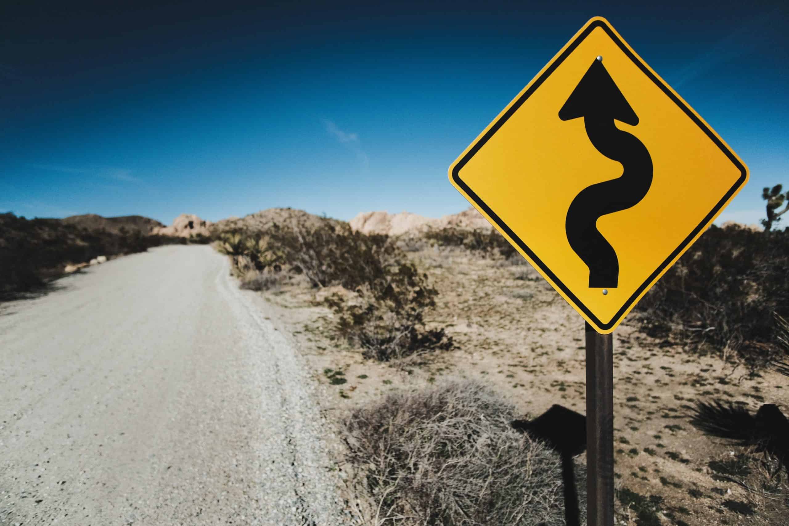 a windy road sign is in the foreground with a gravel road and a desert landscape in the background